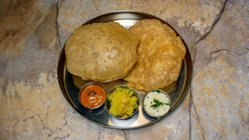 Two golden-brown puri breads served on a steel plate with three small bowls of chutneys.