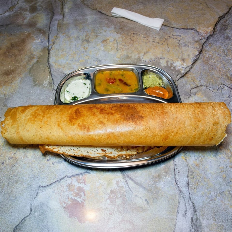 Golden-brown dosa served on a metal plate with four small bowls of chutneys and sambar.