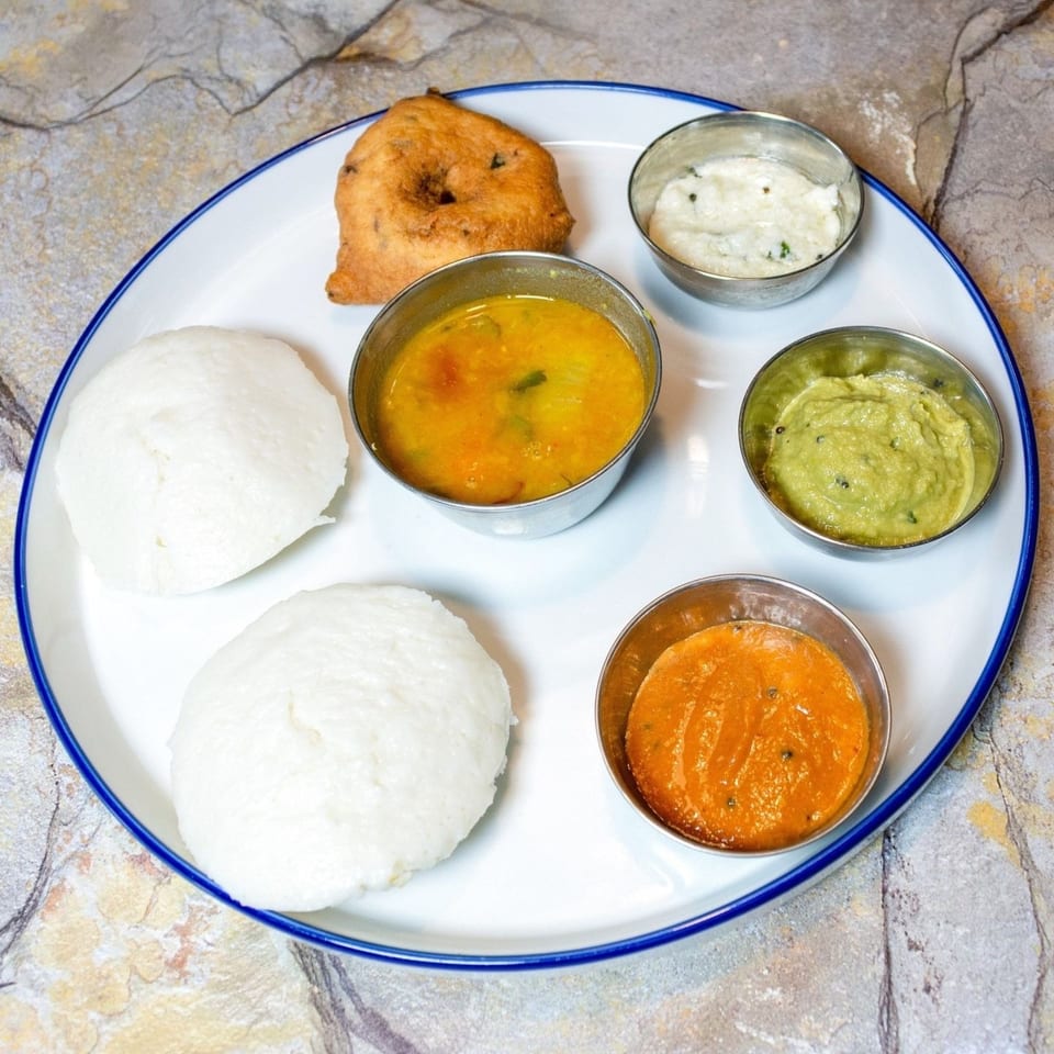 A white plate with two idlis, a vada, sambar, and three chutneys on a stone surface.