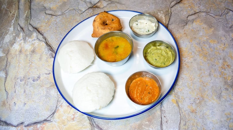 A white plate with two idlis, a vada, sambar, and three chutneys on a stone surface.