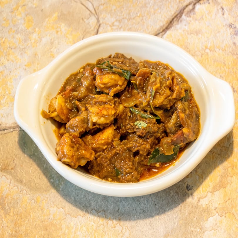 A white bowl filled with a rich, brown curry, garnished with green leaves, on a textured surface.